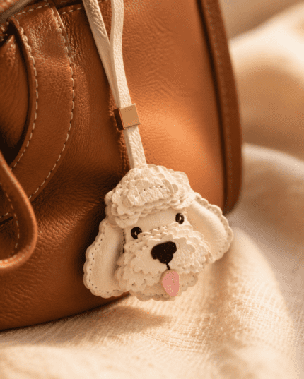 Close-up of handmade white poodle leather bag charm with layered fur, black nose, pink tongue, and gold accent