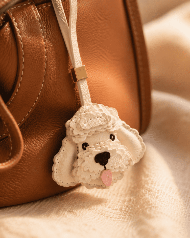 Close-up of handmade white poodle leather bag charm with layered fur, black nose, pink tongue, and gold accent