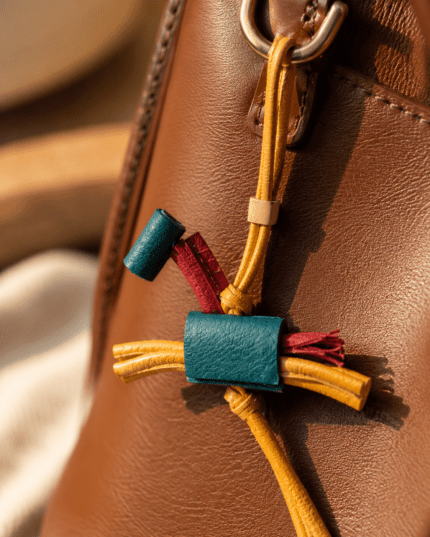 Close-up of handmade colorful leather tassel bag charm with teal blue leather, burgundy accents, mustard yellow strap, cream slider, and gold ring