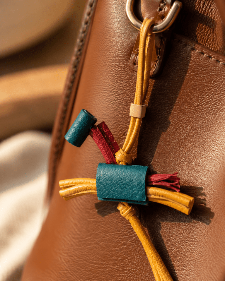 Close-up of handmade colorful leather tassel bag charm with teal blue leather, burgundy accents, mustard yellow strap, cream slider, and gold ring