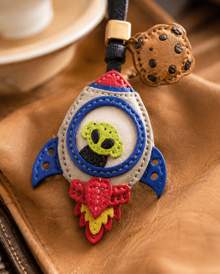 Close-up of handmade rocket alien leather bag charm with green alien face, blue fins, red flame, cookie charm, black strap, and gold slider