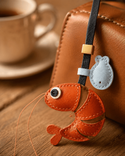 Close-up of handmade shrimp leather bag charm with orange body, long antennae, blue accent charm, black strap, and gold slider