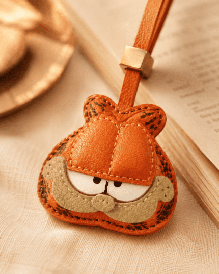 Macro photo of handmade orange cat leather bag charm with sleepy eyes, beige muzzle, orange strap, gold slider, and stitching