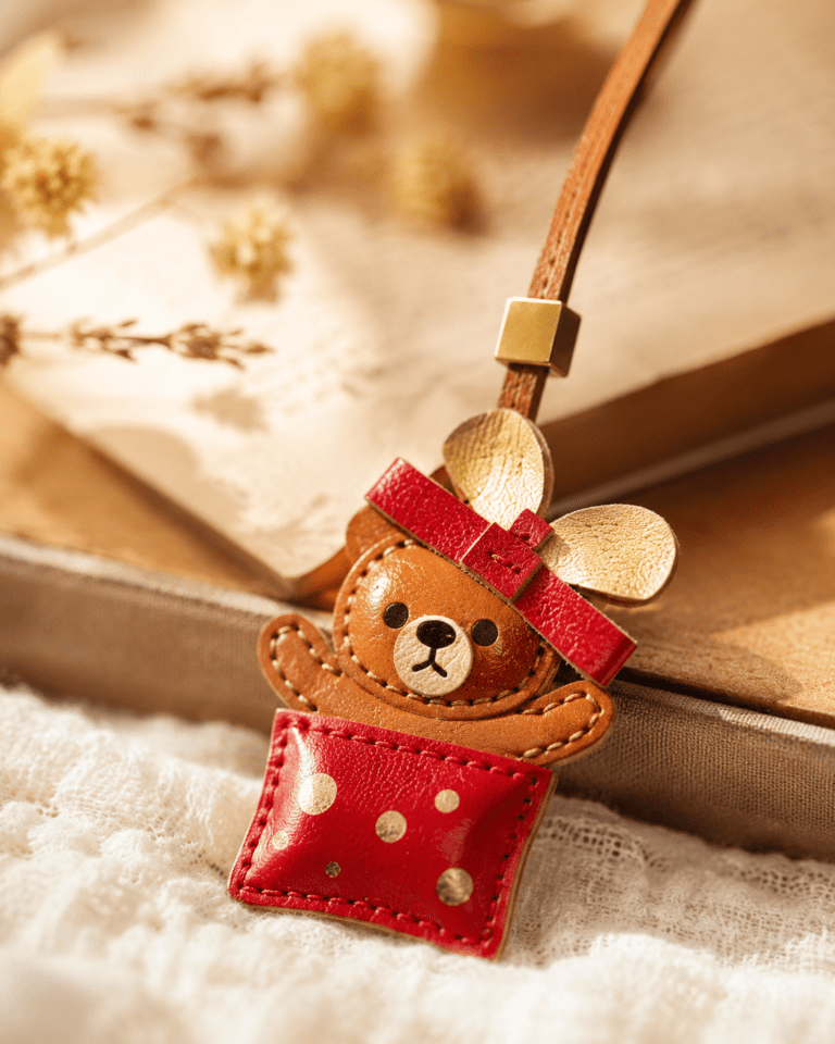 Macro photo of handmade teddy bear leather bag charm with red gift-box pocket, cream ears, brown strap, gold slider, and stitching