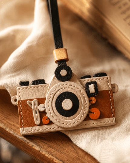 Macro photo of handmade camera leather bag charm with round lens, black strap, gold slider, brown panels, and stitching