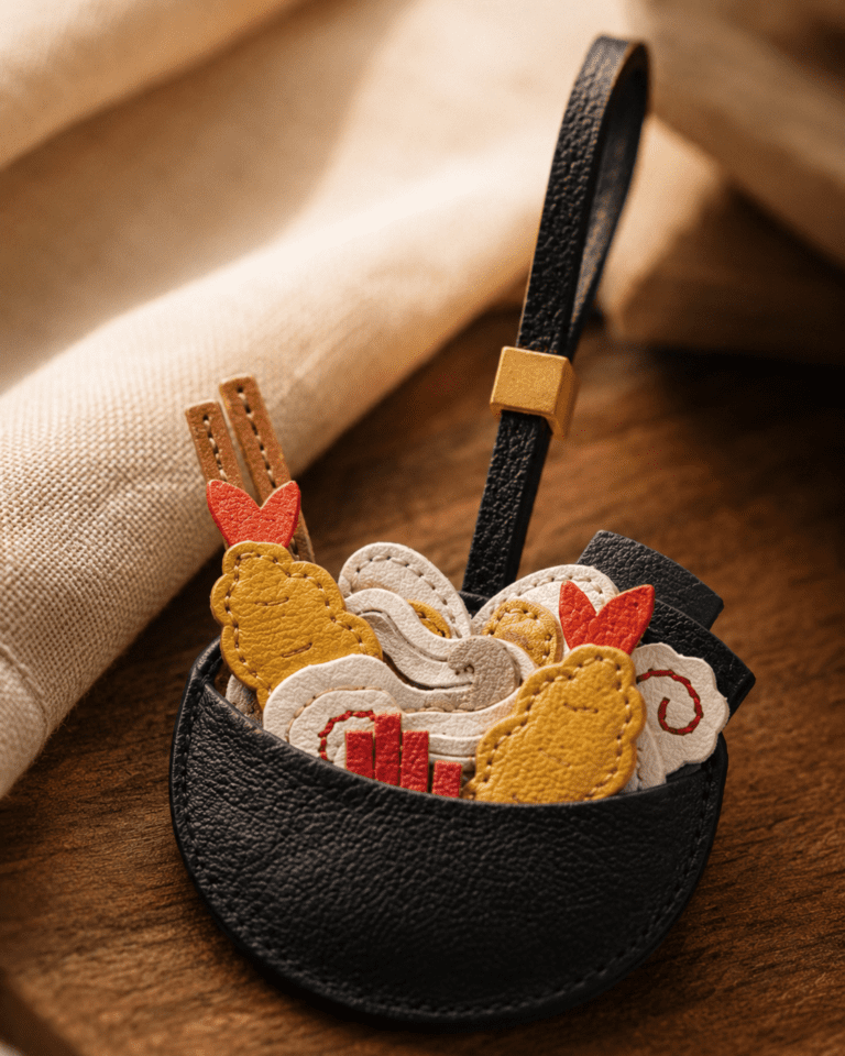 Macro photo of handmade ramen bowl leather bag charm with black bowl, colorful toppings, black strap, and gold slider
