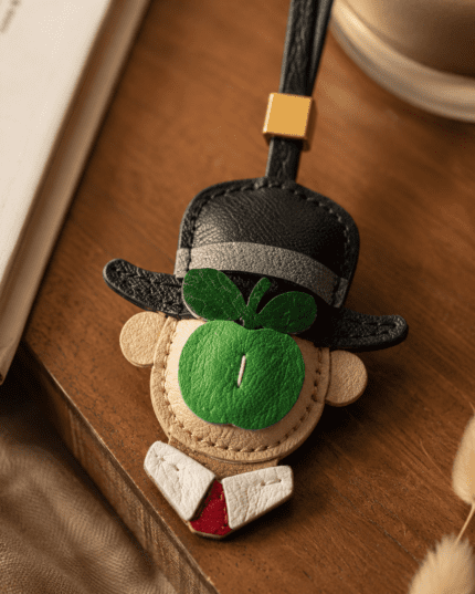 Macro photo of handmade green apple gentleman leather bag charm with black hat, green apple detail, white collar, red tie, black strap, and gold slider