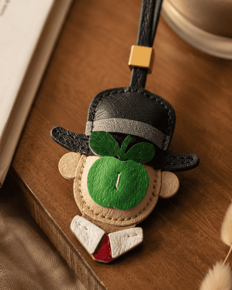 Macro photo of handmade green apple gentleman leather bag charm with black hat, green apple detail, white collar, red tie, black strap, and gold slider