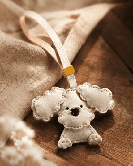 Macro photo of handmade white dog leather bag charm with fluffy ears, black nose, white loop, gold slider, and stitched edges