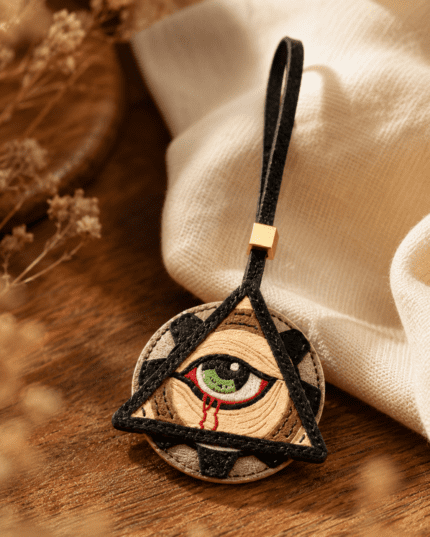Macro photo of handmade mystic eye leather bag charm with green eye, black triangle frame, black loop, gold slider, and stitched leather texture