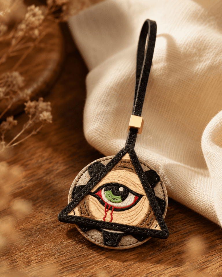 Macro photo of handmade mystic eye leather bag charm with green eye, black triangle frame, black loop, gold slider, and stitched leather texture