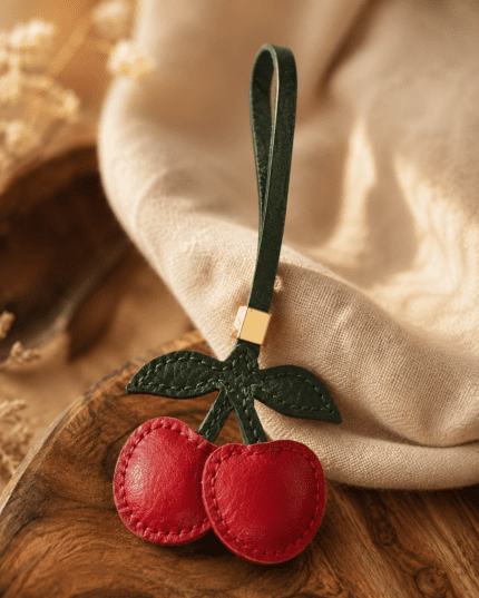 Macro photo of handmade cherry leather bag charm with red cherries, green leaves, green loop, gold slider, and stitched leather texture