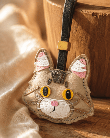 Macro photo of handmade cat leather bag charm with beige leather face, yellow eyes, pink nose, black loop, gold slider, and stitched texture