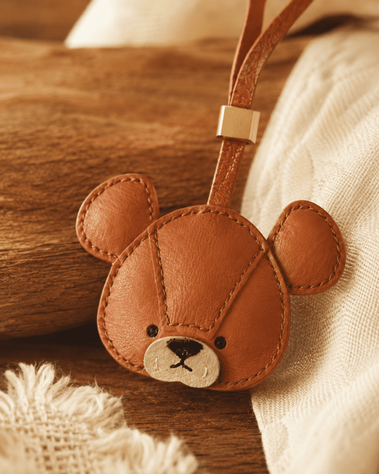 Macro photo of handmade teddy bear leather bag charm with caramel leather face, round ears, beige muzzle, brown loop, gold slider, and stitched texture
