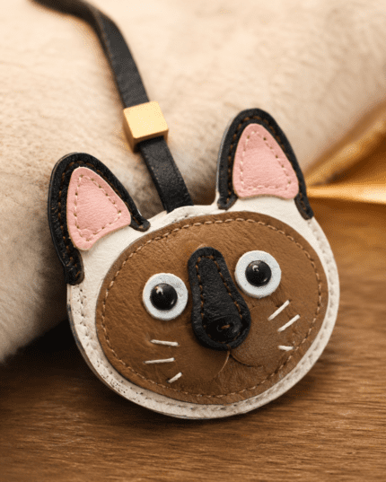 Macro photo of handmade leather cat bag charm with brown cat face, pink ears, black nose, white whiskers, black strap, and gold slider
