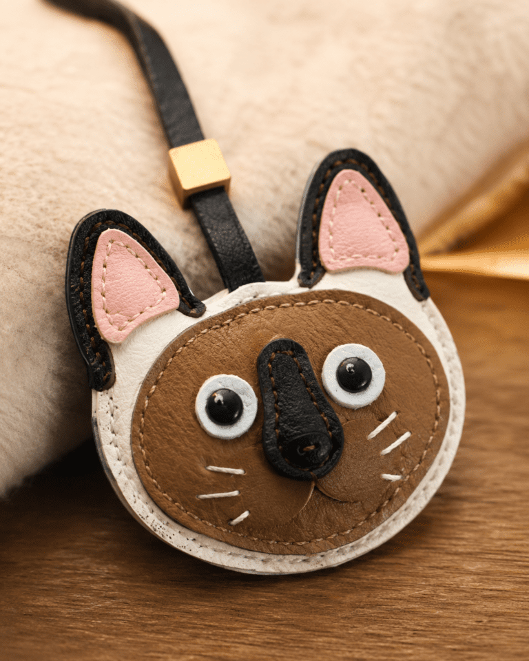 Macro photo of handmade leather cat bag charm with brown cat face, pink ears, black nose, white whiskers, black strap, and gold slider