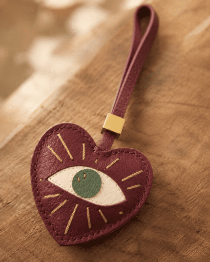 Macro photo of handmade leather heart eye bag charm with burgundy leather, green iris, gold stitching, strap, and gold slider