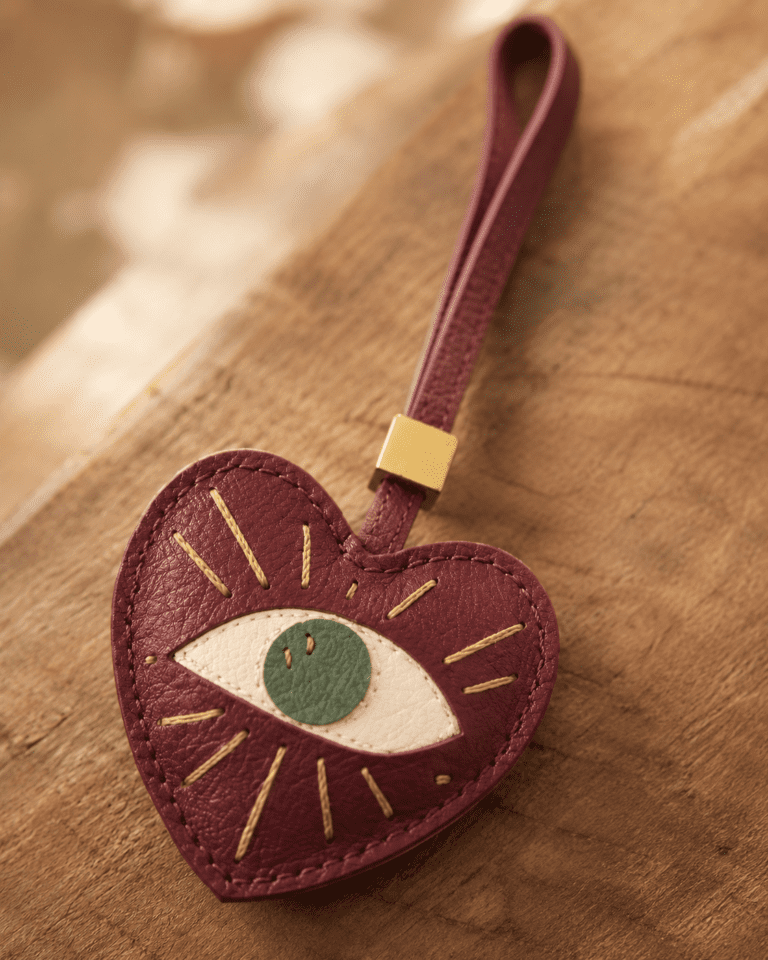 Macro photo of handmade leather heart eye bag charm with burgundy leather, green iris, gold stitching, strap, and gold slider