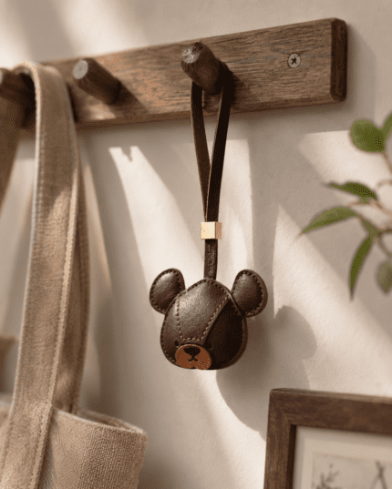 Handmade dark brown leather bear bag charm hanging from a wooden wall hook in warm sunlight