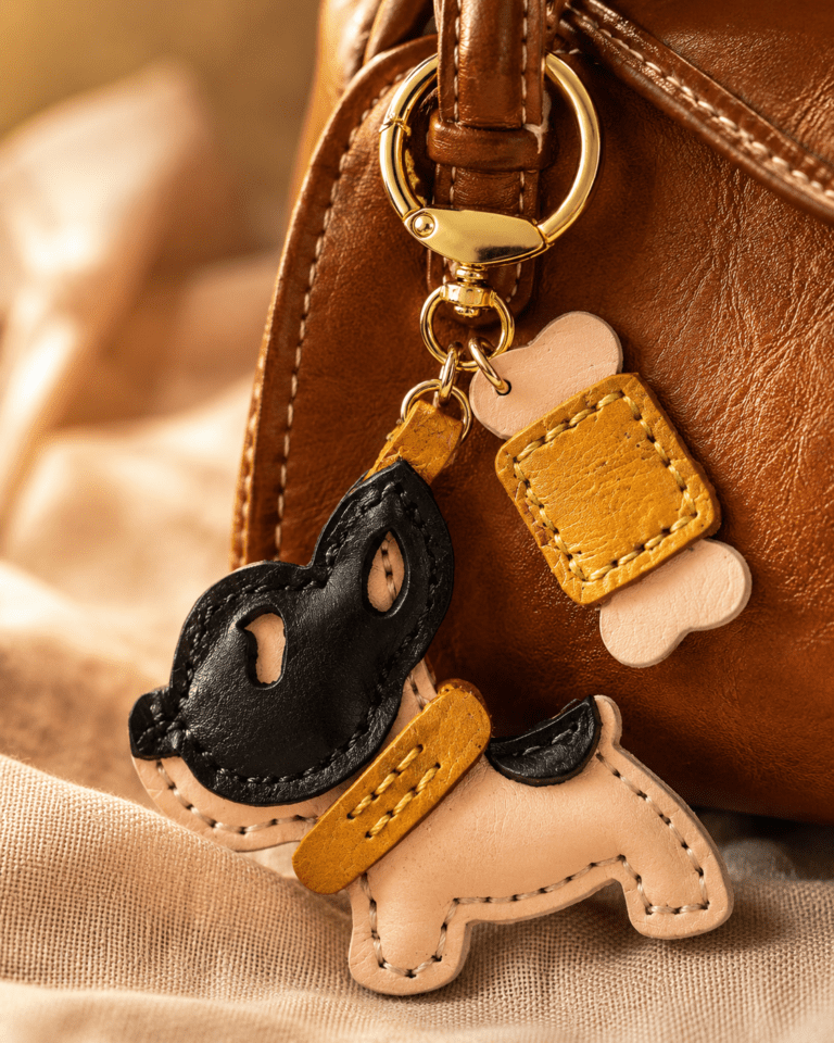 Close-up of a handmade leather black mask puppy bag charm with bone accent and gold clasp on a brown handbag