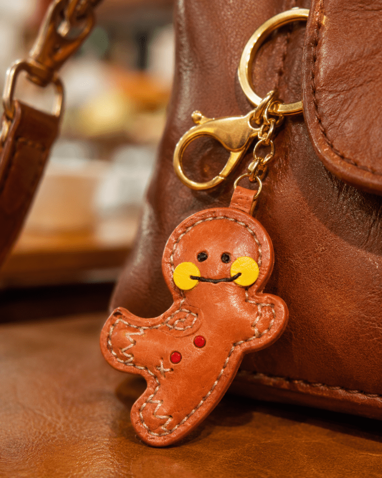 Close-up of a handmade leather gingerbread man bag charm with yellow cheeks, red buttons, white stitching, and gold clasp