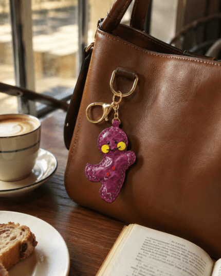 Handmade leather magenta gingerbread man bag charm hanging on a brown leather handbag in a warm cafe scene