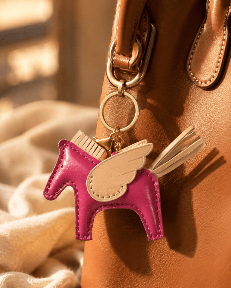 Close-up of handmade magenta leather winged horse bag charm with cream wing, beige tail, pink stitching, and gold hardware
