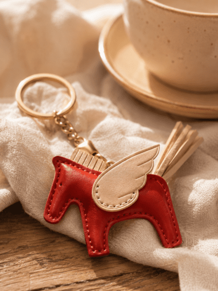 Close-up of handmade red leather winged horse bag charm with cream wing, pale tail, stitching, and gold hardware