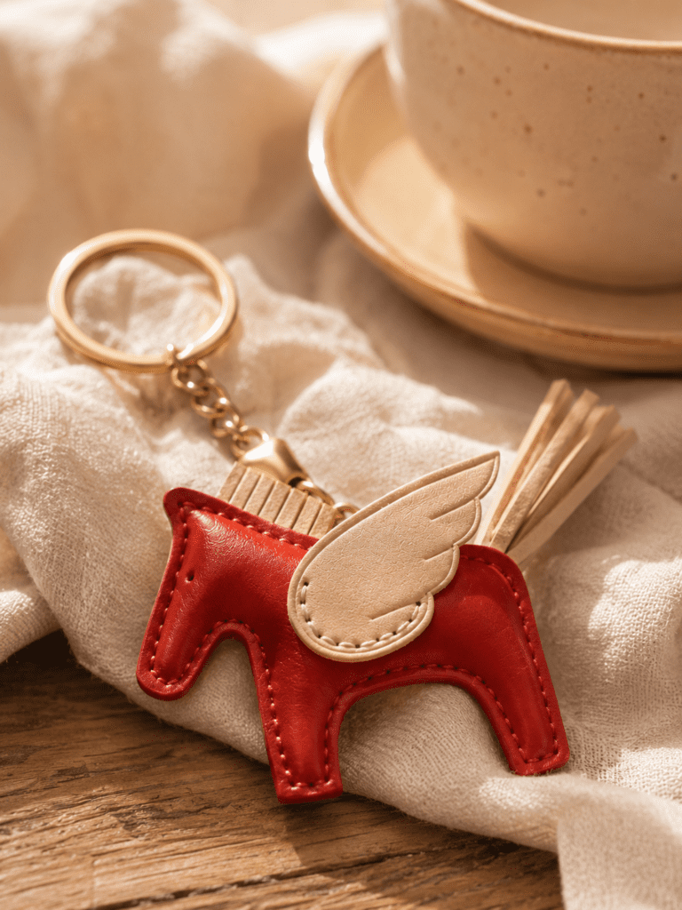 Close-up of handmade red leather winged horse bag charm with cream wing, pale tail, stitching, and gold hardware