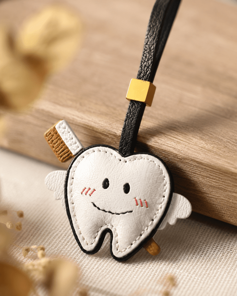 Close-up of handmade tooth leather charm with black strap, gold slider, wings, toothbrush detail, and smiling face