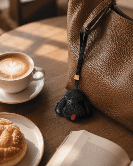 Handmade leather black poodle charm beside a brown leather bag with coffee, pastry, and book in warm cafe light