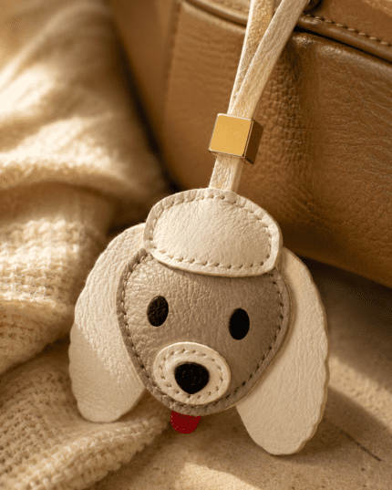 Close-up of handmade leather dog charm with cream ears, beige face, gold accent, and textured stitching.