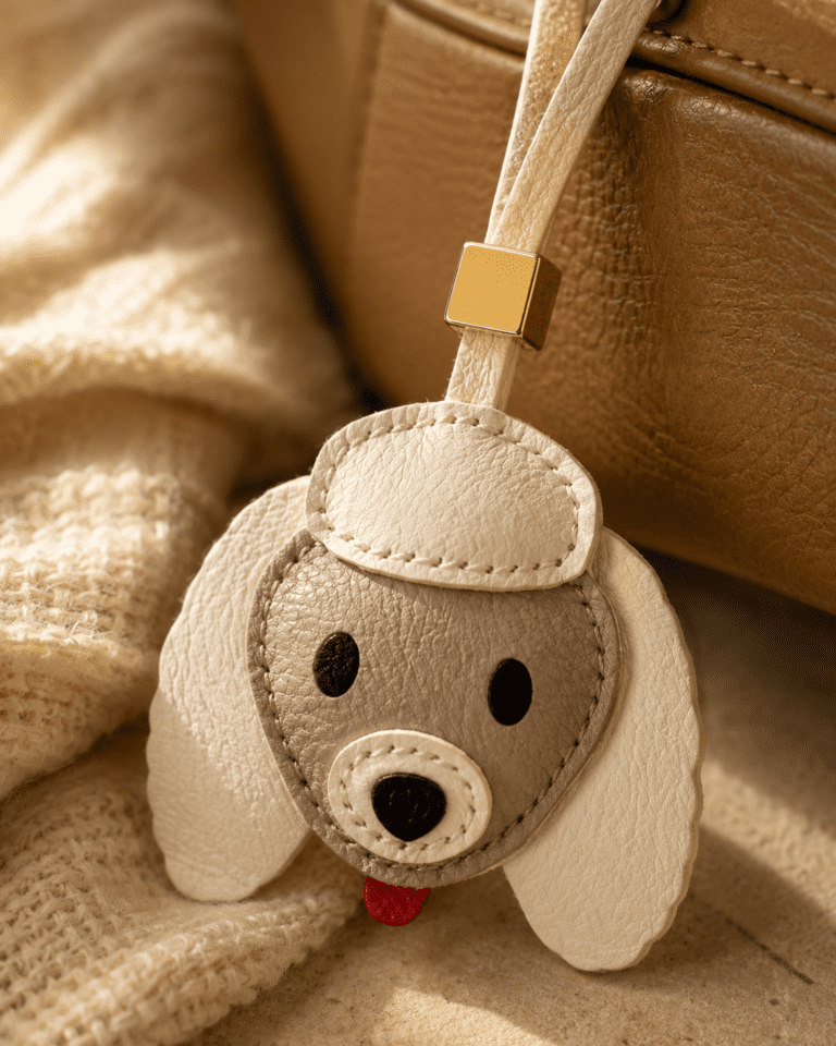 Close-up of handmade leather dog charm with cream ears, beige face, gold accent, and textured stitching.