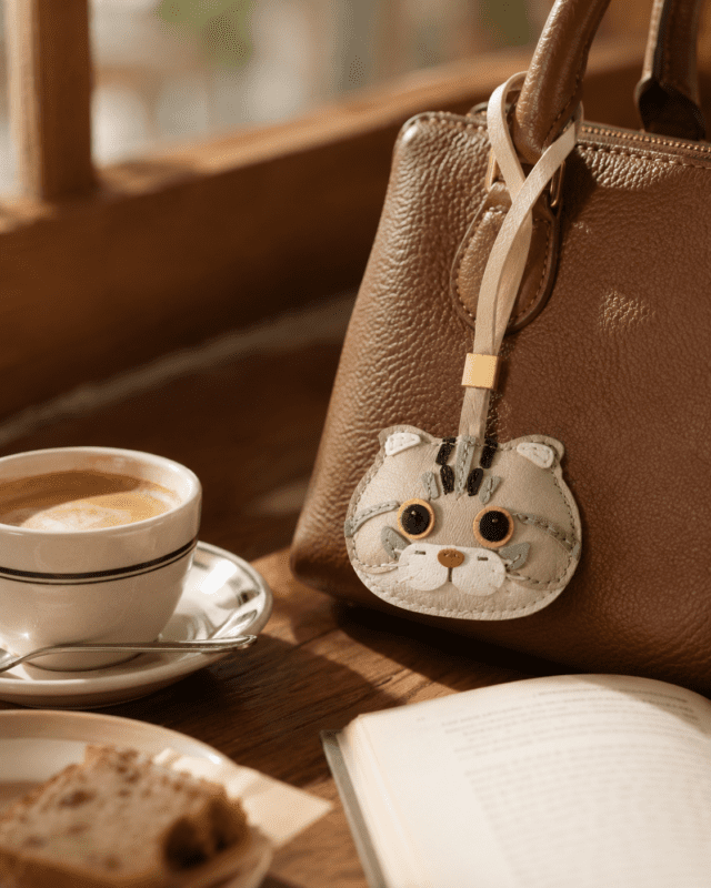 Alt Text: Handmade leather tabby cat head charm attached to brown leather handbag, lifestyle coffee scene