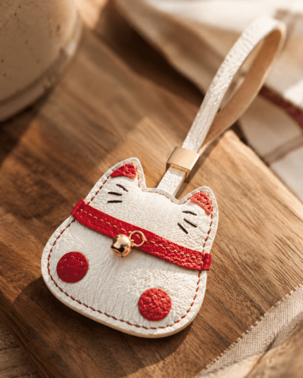 Close-up of a handmade white lucky cat leather charm with red collar and gold bell on a wooden surface.
