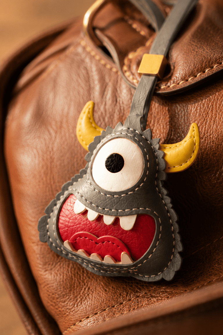Close-up of a handmade gray one-eyed monster leather bag charm showing yellow horns, red mouth, and stitched details.