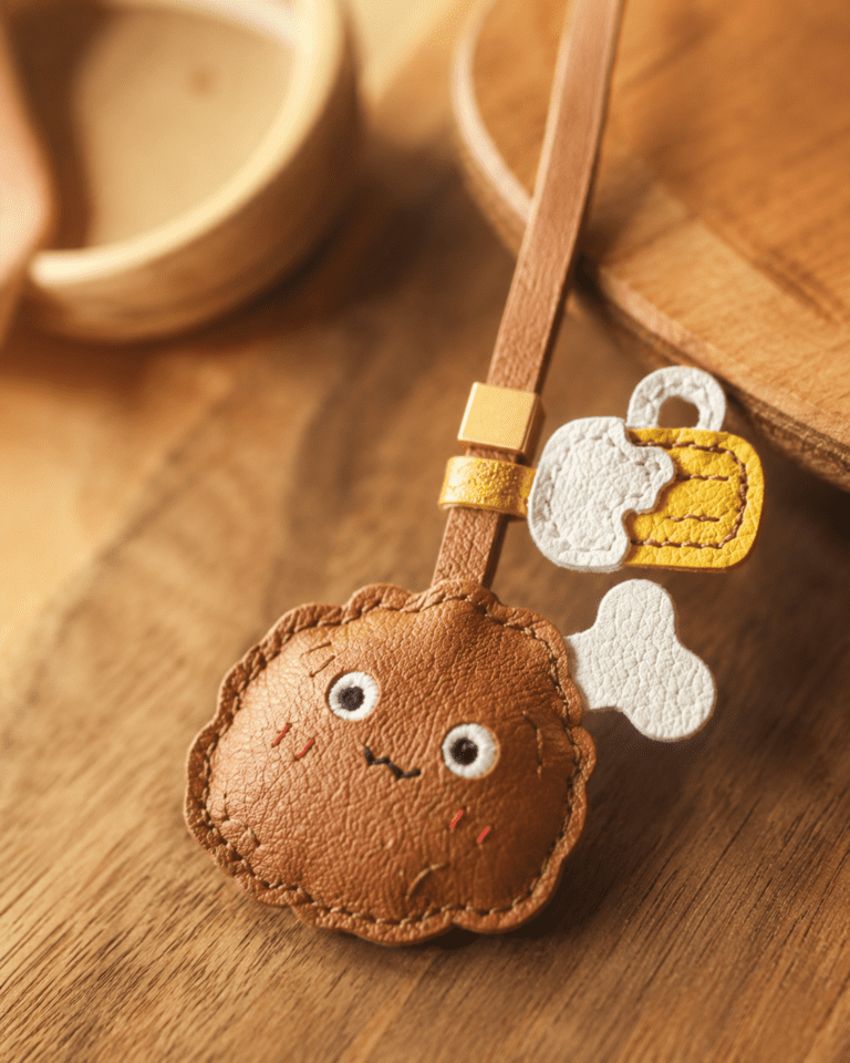 Close-up of handmade leather fried chicken and beer bag charm showing brown leather texture, cute face, white bone, yellow beer detail, stitching, and gold slider.