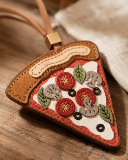 Close-up of a handmade leather pizza slice bag charm showing red tomato slices, green leaves, mushrooms, black olives, stitched crust, strap, and gold slider.