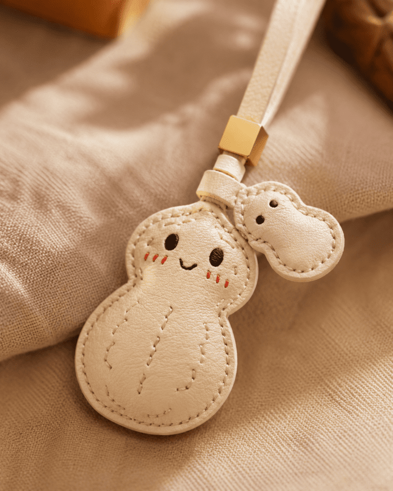 Close-up of a handmade cream ghost leather bag charm showing stitched edges, smiling face, blush details, white strap, and gold slider.